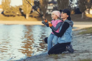 A woman teaches a child to fish by a lake on a sunny day.